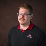 Alt text: A man with short light brown hair, a mustache, and glasses smiles while looking slightly to the side. He is wearing a black sweater with a red collared shirt underneath. The sweater has the Chico State logo embroidered on it. The background is a dark, neutral gradient.